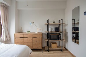 Een slaapkamer met een houten dressoir, een nachtkastje met een lamp, een plank met een waterkoker en een magnetron, en een grote spiegel aan de muur.