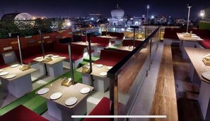 Ein Restaurant auf dem Dach mit mehreren Tischen und Stühlen, im Hintergrund eine Stadtansicht und ein klarer Nachthimmel.