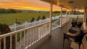 En overdækket veranda med udsigt til solnedgang over en græsplæne, hvidt stakerværk og fletningsmøbler.