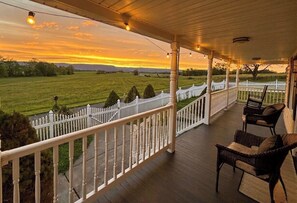 Un porche cubierto con vistas a una puesta de sol sobre un campo verde, cerca de una valla de piquetes blanca y con muebles de mimbre.