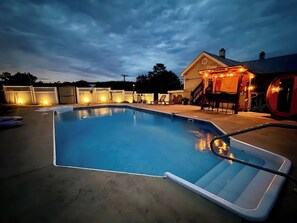 Una piscina rectangular con una escalera, rodeada por una cerca, y un área de patio techada con luces.