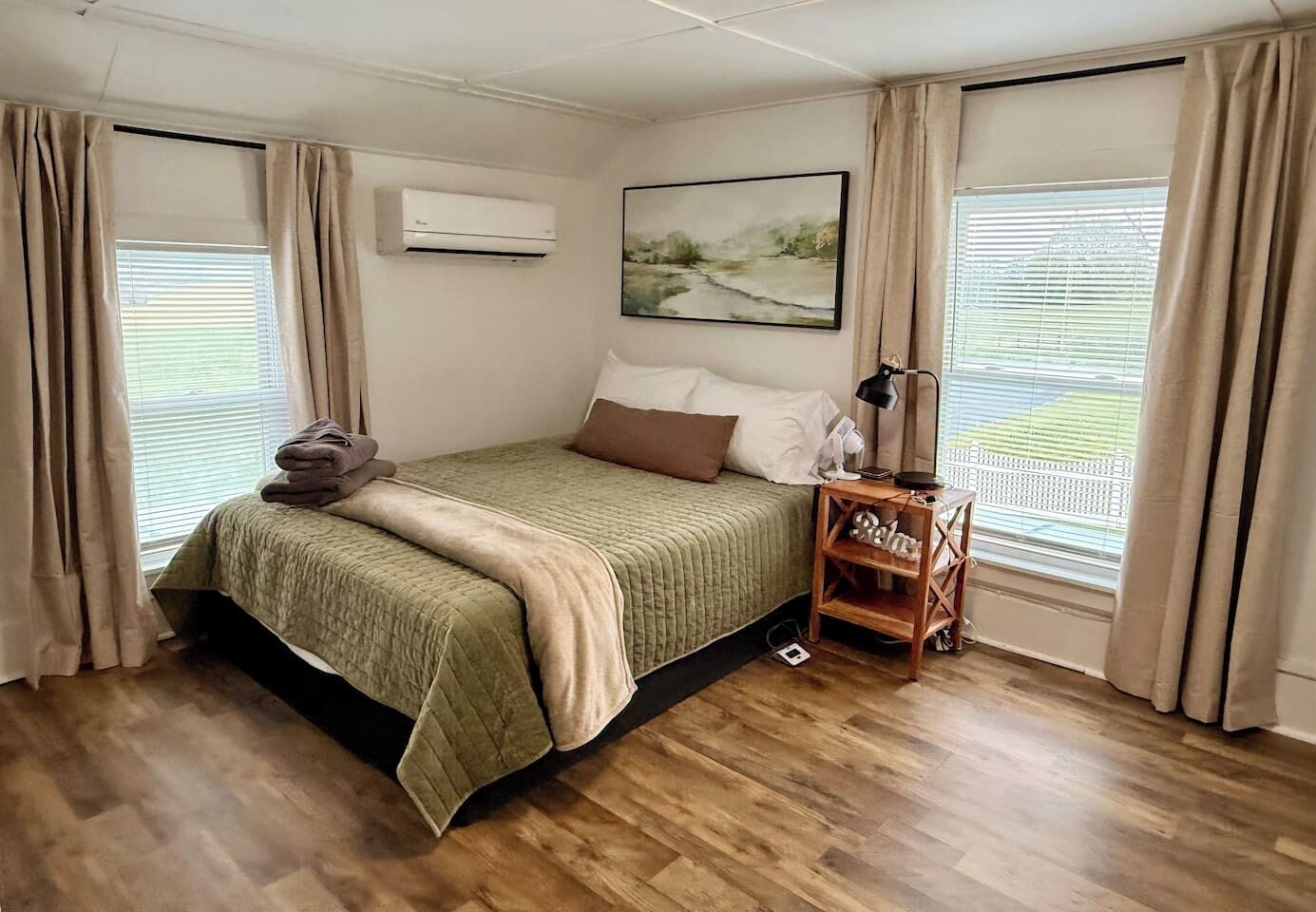 A bedroom with a bed, a nightstand, an air conditioner, a window with blinds, and a painting on the wall.