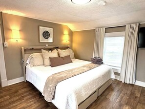 Un dormitorio con cama, lámparas de noche, un cuadro enmarcado, ventana con cortinas y televisión en la pared.