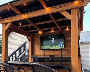 Una pérgola de madera con luces colgantes, una pantalla de televisión mostrando un evento deportivo y mobiliario de exterior.