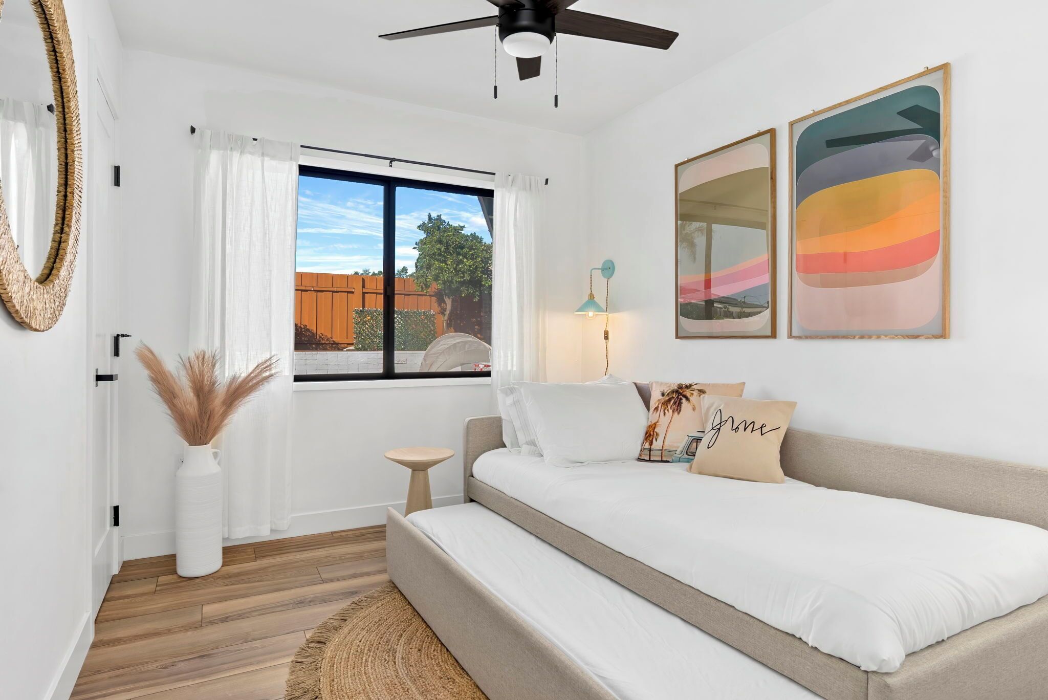 Un dormitorio con cama, ventilador de techo, ventana con cortinas, mesita de noche, lámpara y dos cuadros abstractos.