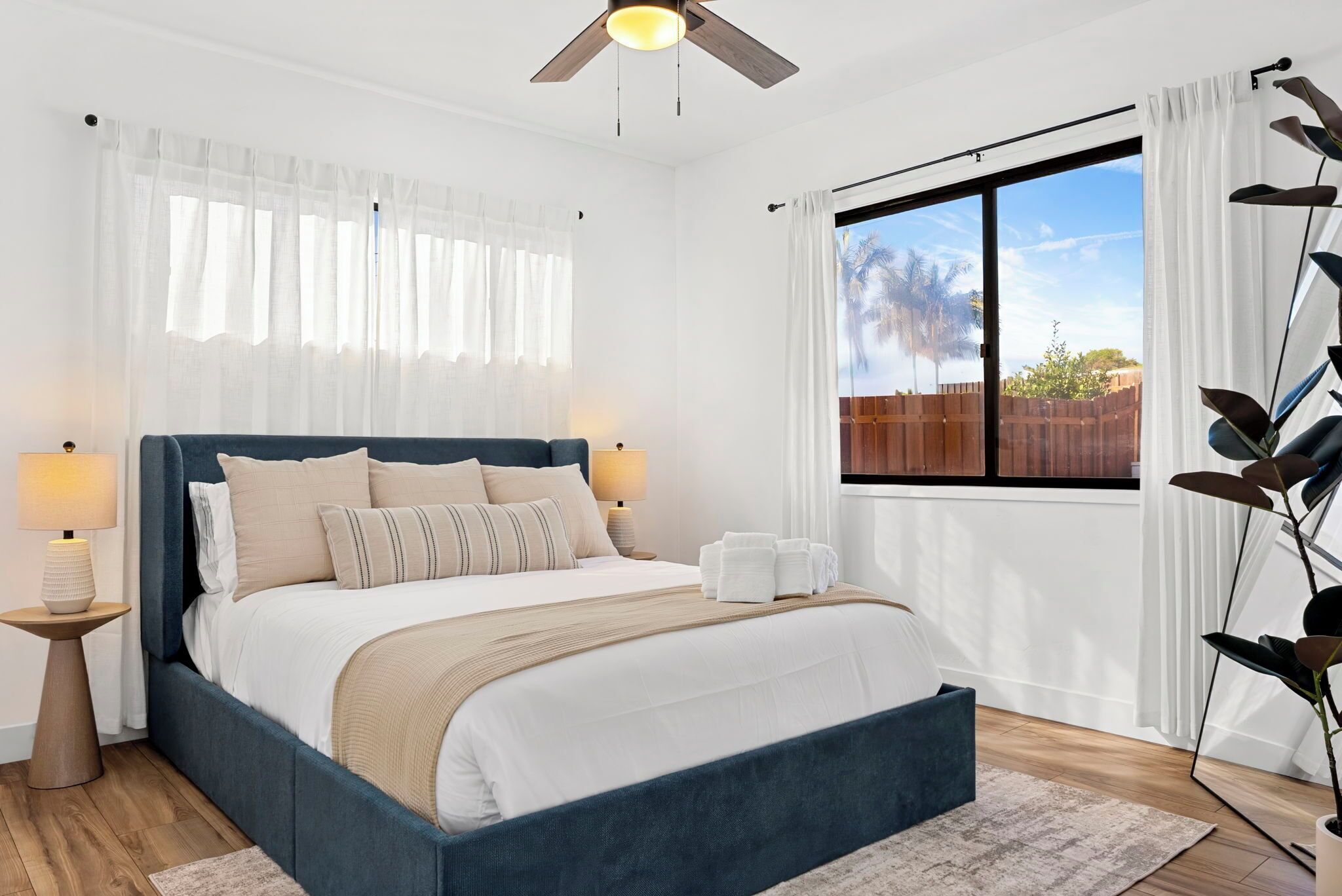 A bedroom with a large bed, two bedside lamps, a ceiling fan, a window with curtains, and a view of the outdoors.