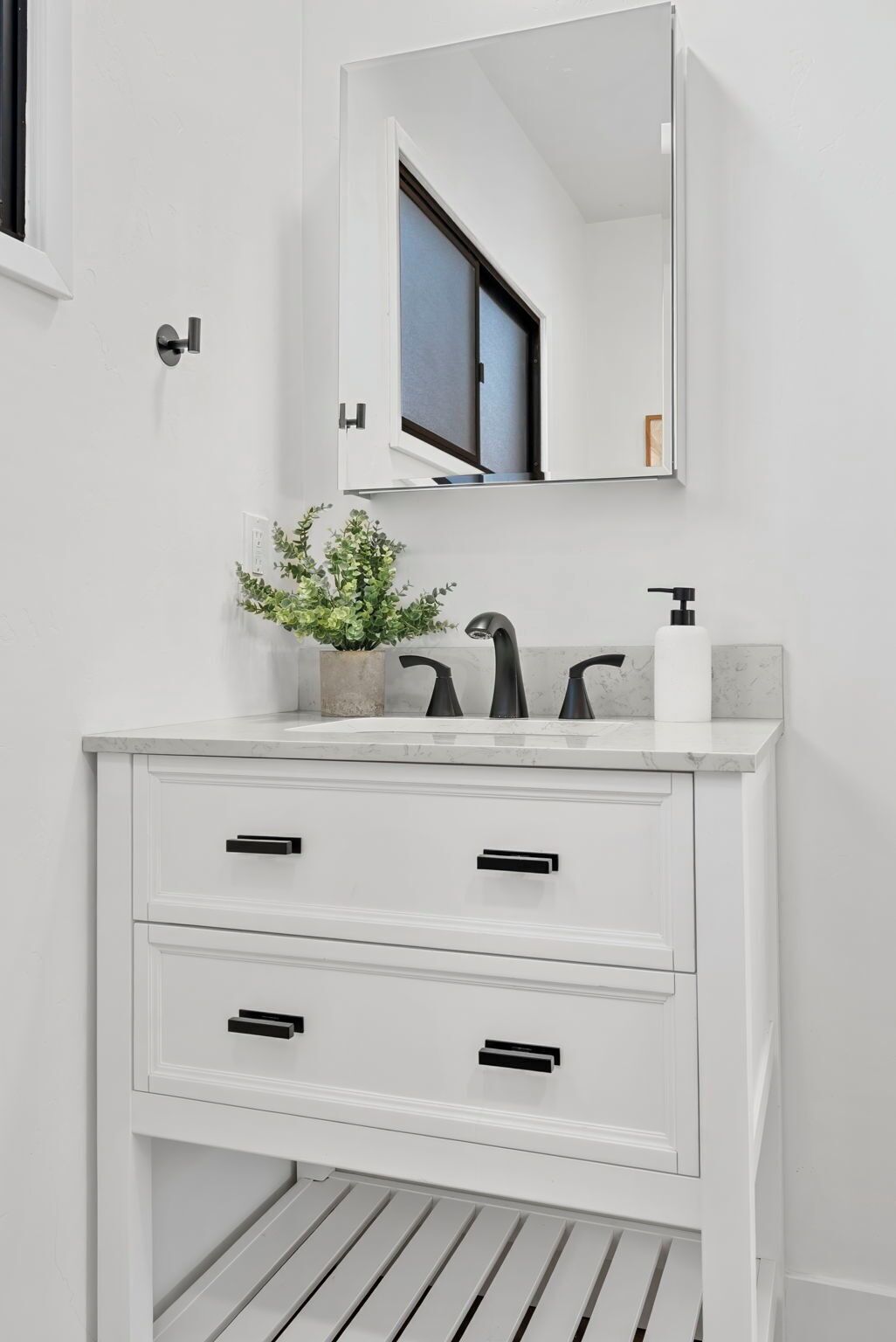 A white vanity with a mirror, three black handles, and a small potted plant.