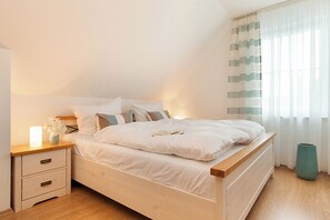 A bedroom with a wooden bed, white bedding, a bedside table with drawers, a vase with flowers, and a window with sheer curtains. - Detached vacation home│3 bedrooms│2 bathrooms │Whirlpool│Sauna│WLAN (Zingst)