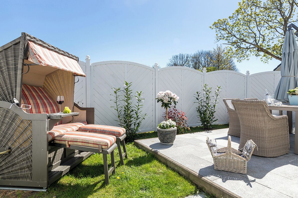Ein Patio im Freien mit Korbmöbeln, einer gepolsterten Bank, einem Tisch mit einem Weinglas und Kübelpflanzen.