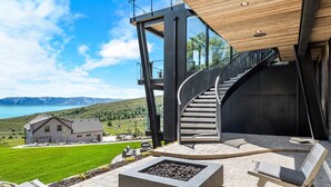 Un patio moderno al aire libre con un hogar para fuego, asientos y vistas a un lago y montañas.