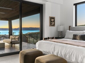 A bedroom with a large bed, a nightstand, a lamp, a chair, and a view of the ocean.