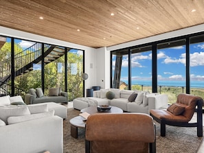 A spacious living area with a modern design, featuring a staircase, large windows, and a view of the outdoors.