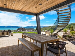 An outdoor patio with a wooden table and benches, a spiral staircase, and a view of mountains and a lake.