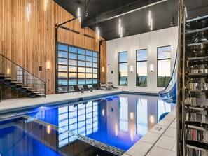 An indoor swimming pool with a view of mountains through large windows, a staircase, and a wooden wall.
