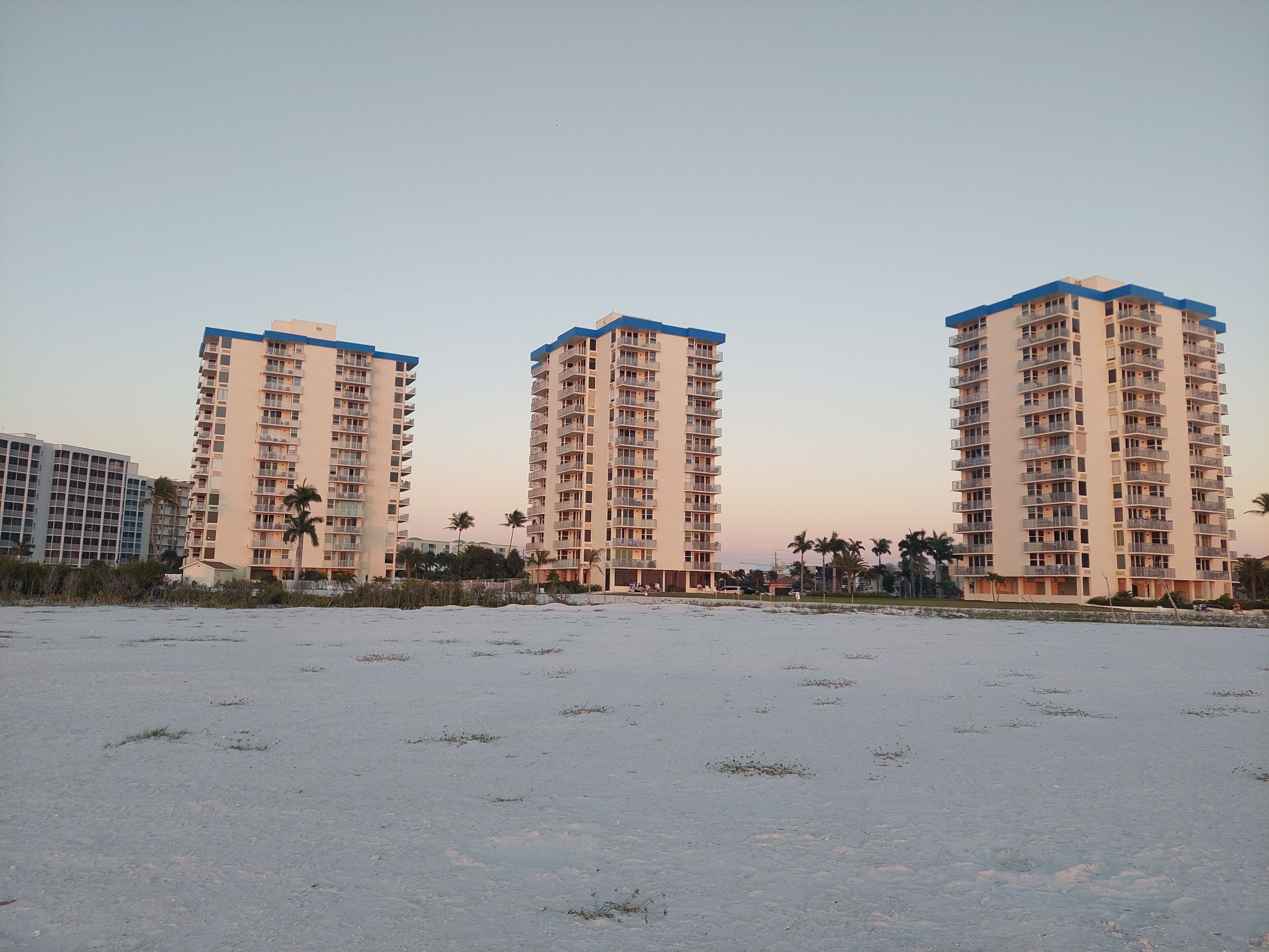 Trois immeubles résidentiels de grande hauteur, dotés de balcons, sont situés sur une plage de sable où la végétation est clairsemée.