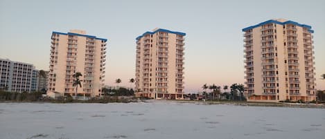Trois immeubles résidentiels de grande hauteur, dotés de balcons, sont situés sur une plage de sable où la végétation est clairsemée.