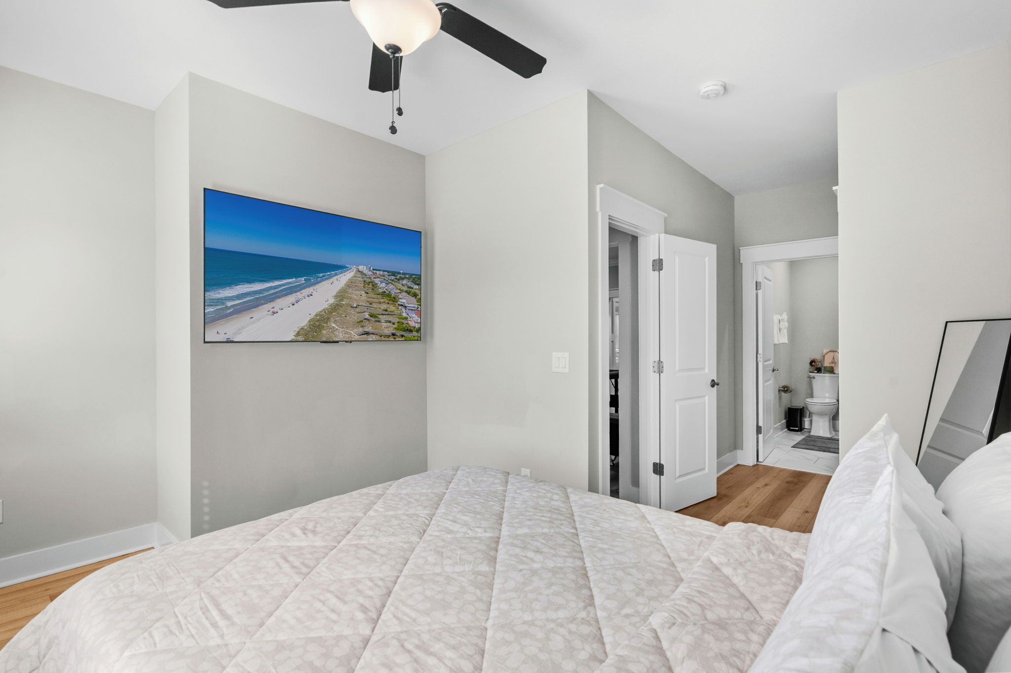 A bedroom with a bed, a ceiling fan, a painting of a beach, and a door leading to a bathroom.