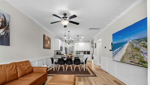En moderne stue med en brun lædersofa, et spiseområde med bord og stole, en loftsvifte og et stort tv, der viser et strandmotiv.