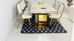 Un espace repas avec une table blanche, des chaises dorées et un tapis à motifs.