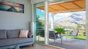 A modern living room with a grey sofa, a glass coffee table, and a view of a mountainous landscape through large sliding glass doors. - Villa dei Sospiri 4, Emma Villas (Castellammare del Golfo)