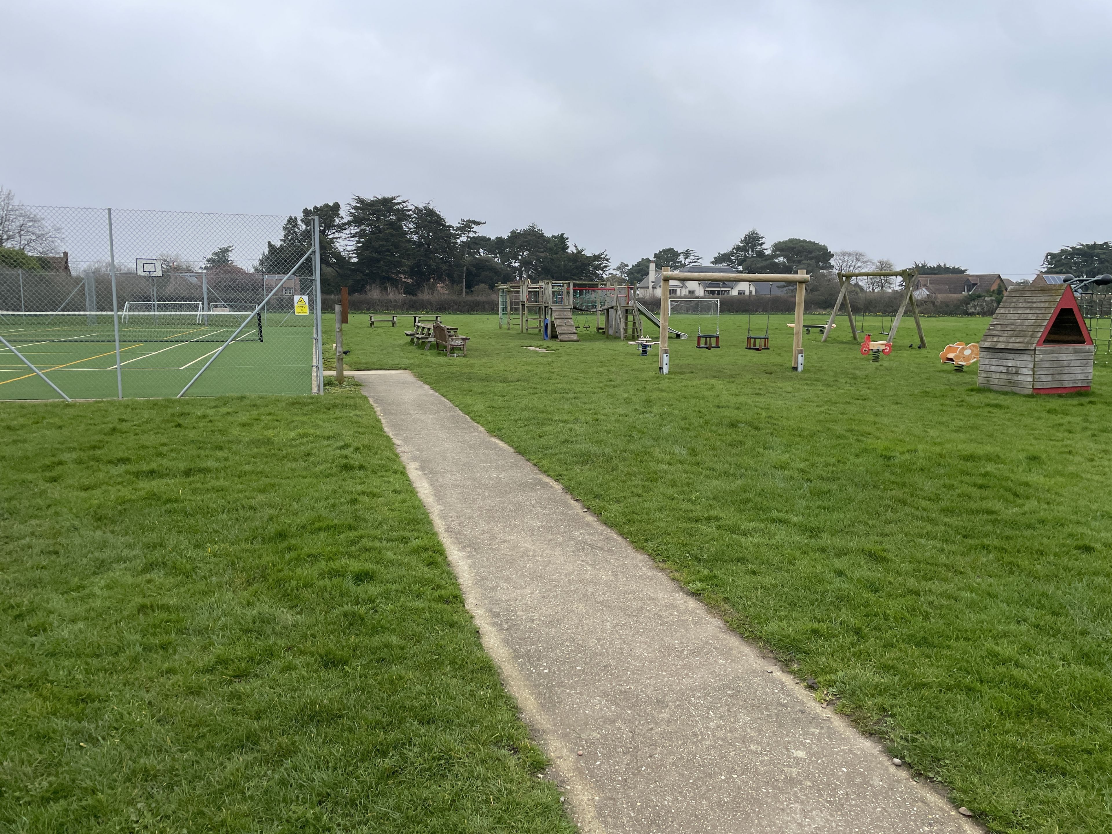 Un campo cubierto de césped con un camino pavimentado, juegos infantiles y una zona delimitada por una valla.