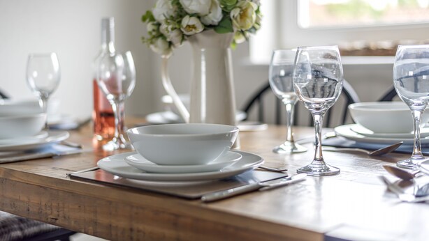 Una mesa de comedor dispuesta con platos, tazones y copas de vino blancos, un florero con flores blancas y una botella de vino rosado.