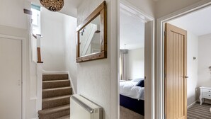 Un pasillo con escaleras cubiertas de alfombra, un radiador, una puerta de madera y una cama en la habitación contigua.