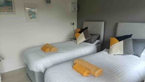 A hotel room with two beds, each with a rolled towel. The room has a neutral color scheme with light walls and a gray headboard.