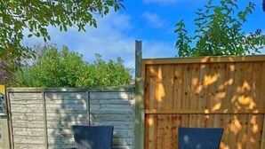 An outdoor seating area with two wicker chairs and a small round table, a potted plant with pink flowers, and a wooden fence in the background.