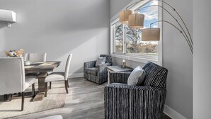 Un coin repas comprenant une table, des chaises et un canapé. Une fenêtre offre une vue sur les arbres, et une lampe est suspendue au-dessus de la table.