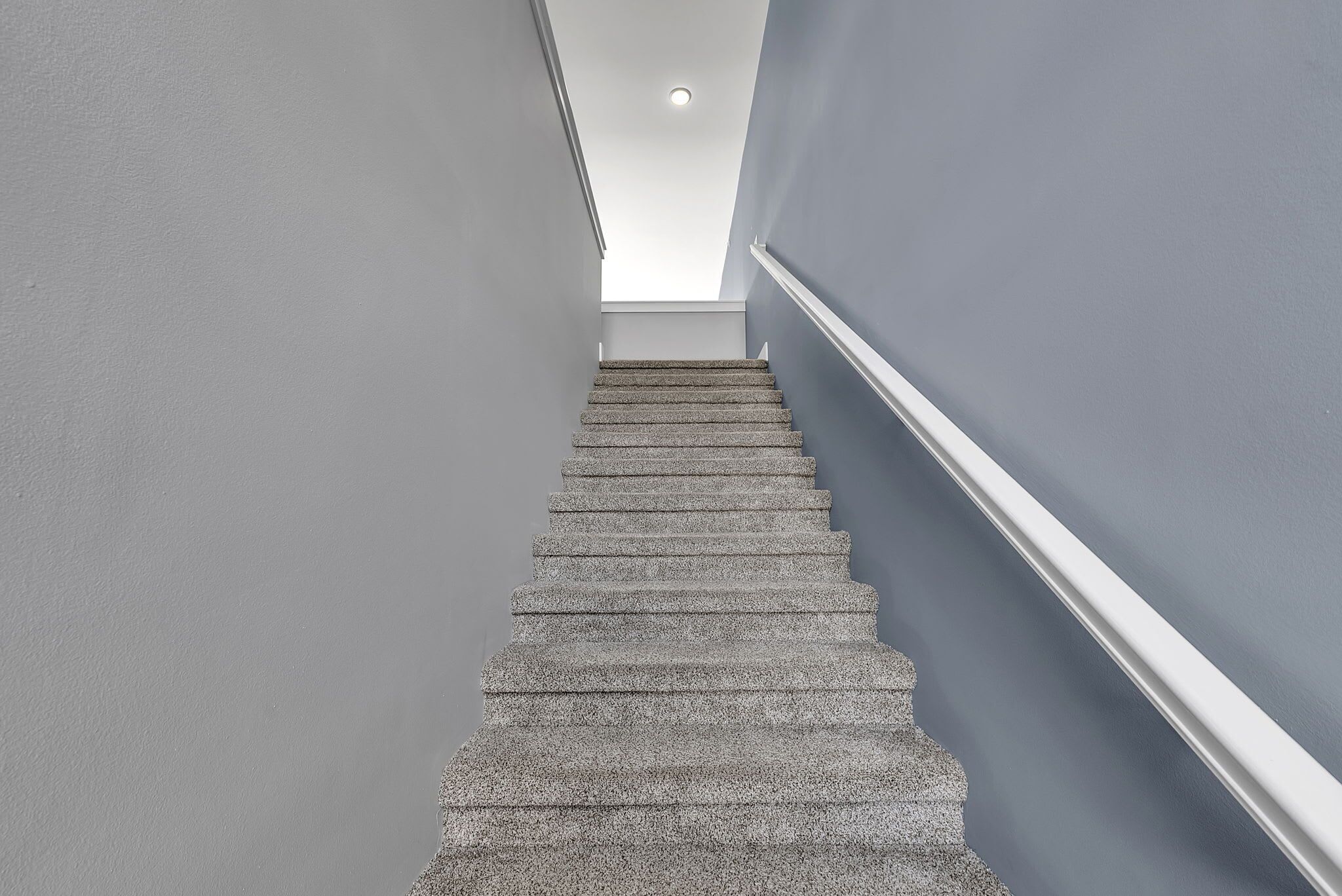 Un escalier muni d’une rampe, menant à un étage où se trouve une pièce avec un plafond blanc et un luminaire apparent.