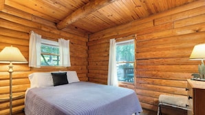A log cabin bedroom with a bed, two windows with curtains, a nightstand, and a lamp. - Table Rock Lake Cabin - The Oaks #5 with Lake Access & Hot Tub (Shell Knob)