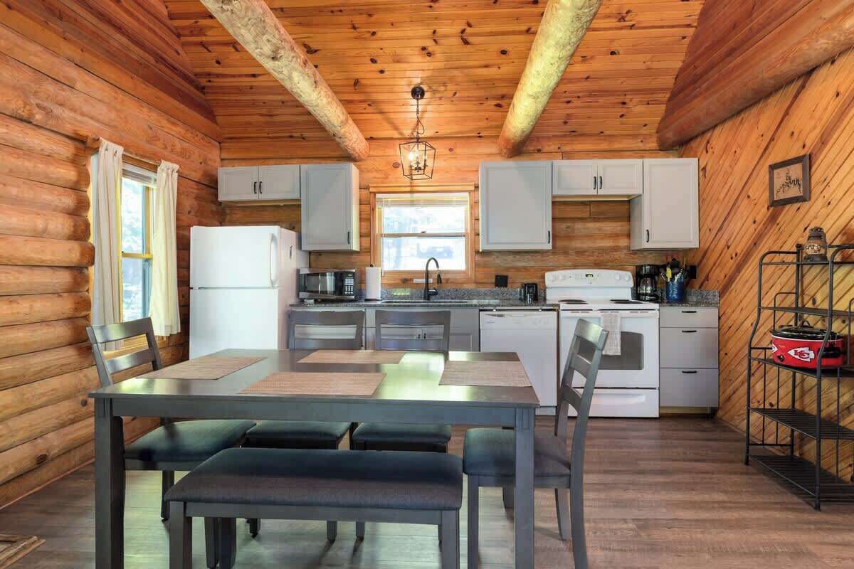 Interior de una cabaña de madera con una mesa de comedor, sillas, una cocina con armarios blancos, un refrigerador y una lámpara colgante.