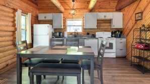 A log cabin interior with a dining table, chairs, a kitchen with white cabinets, a refrigerator, and a hanging light fixture. - Table Rock Lake Cabin - The Oaks #5 with Lake Access & Hot Tub (Shell Knob)