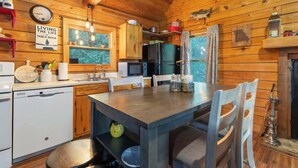 A wooden cabin interior with a kitchen, dining table, chairs, a refrigerator, a microwave, a dishwasher, and a window with a view of trees. - Table Rock Lake Cabin - The Cedars #1 with Lake Access, Hot Tub, Outdoor Pool (Shell Knob)
