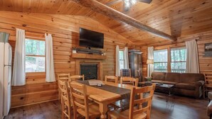 Interno di una baita in legno con tavolo da pranzo, sedie, divano, televisore e camino.