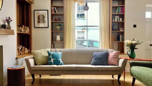 Ein Wohnzimmer mit einem beigefarbenen Sofa, zwei Beistelltischen, einem grünen Sessel und einem großen Fenster mit Vorhängen.