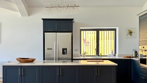 Uma cozinha moderna com ilha central, armários escuros, uma geladeira de aço inoxidável, um bebedouro e uma janela grande com persianas verticais.