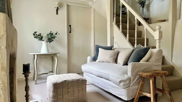Een gezellige woonkamer met een witte bank, een klein houten krukje en een rieten poef.