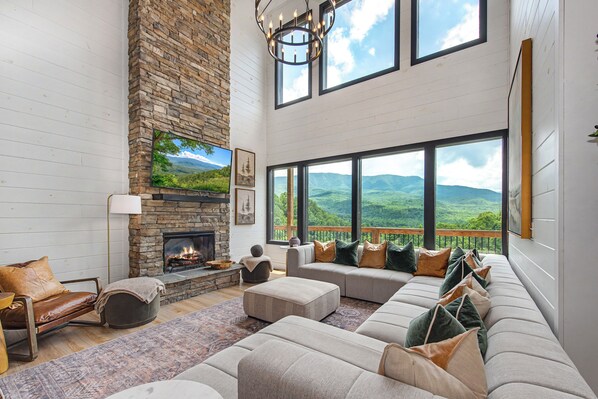 A spacious living room with a stone fireplace, large windows, and a view of the mountains. - Modern Mountain Luxury 4BR 4.5BA with Indoor Pool and Incredible Views (Gatlinburg)