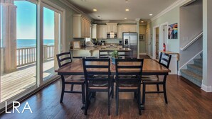 A spacious kitchen with a large island, a dining area with a wooden table and chairs, and a view of the ocean through large windows. - Inn Luxury North | Livin’ the Luxe Life (Surf City)