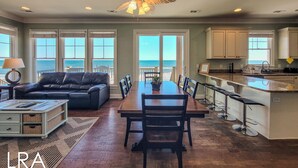 A spacious living room with a large wooden dining table, a black leather sofa, and a view of the ocean through the windows. - Inn Luxury North | Livin’ the Luxe Life (Surf City)