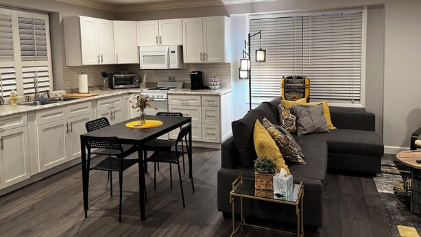 Una cocina moderna con gabinetes blancos, microondas y estufa. Hay un comedor con mesa y sillas negras, y un sofá negro con cojines amarillos.