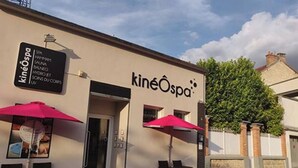 KineOspa-niminen kylpylä, jonka kyltissä luetellaan tarjolla olevat palvelut, kuten SPA-hoidot, hydroterapia ja hieronta. Ulkona on kaksi oleskelualuetta, joissa on aurinkovarjot.