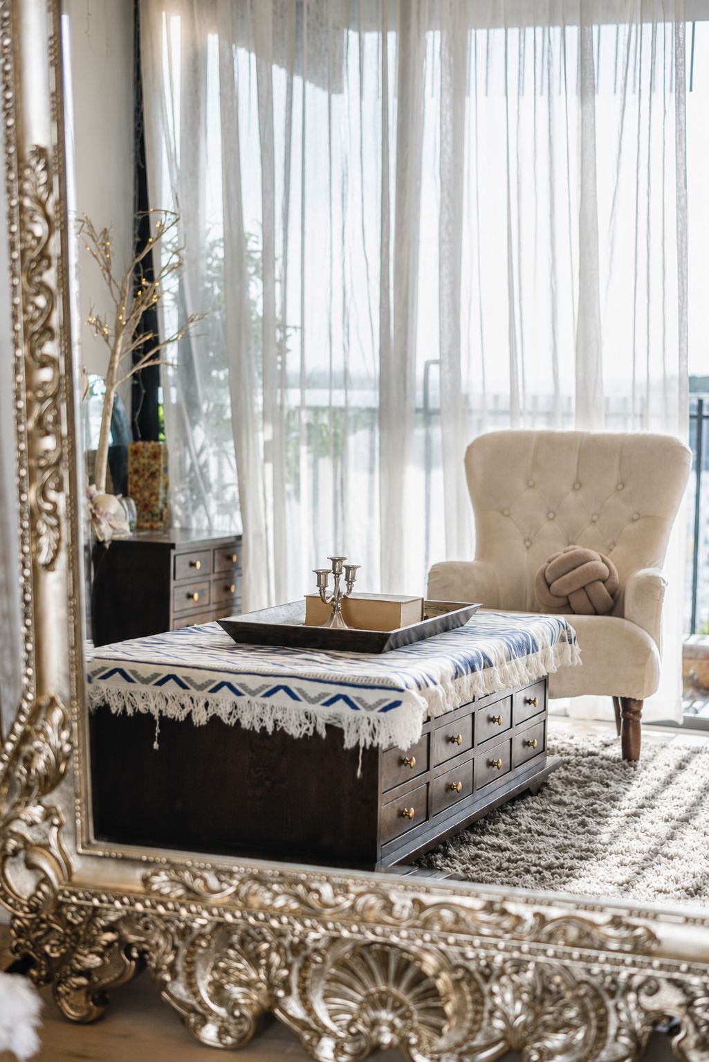 A room with a large ornate mirror, a beige armchair, a wooden chest of drawers, and a patterned throw on a ottoman.
