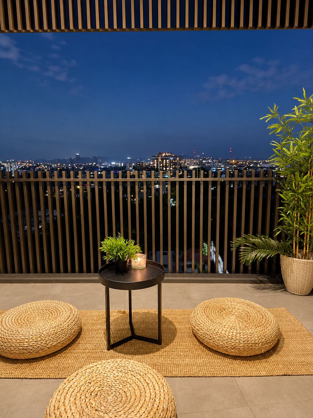 A balcony with a view of a cityscape at dusk, featuring a wooden fence, a small round table with a plant, and two woven poufs.