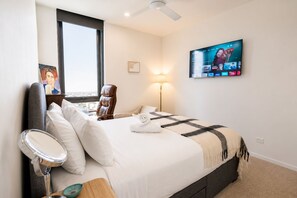 A hotel room with a bed, a chair, a desk with a lamp, a TV mounted on the wall, and a window.