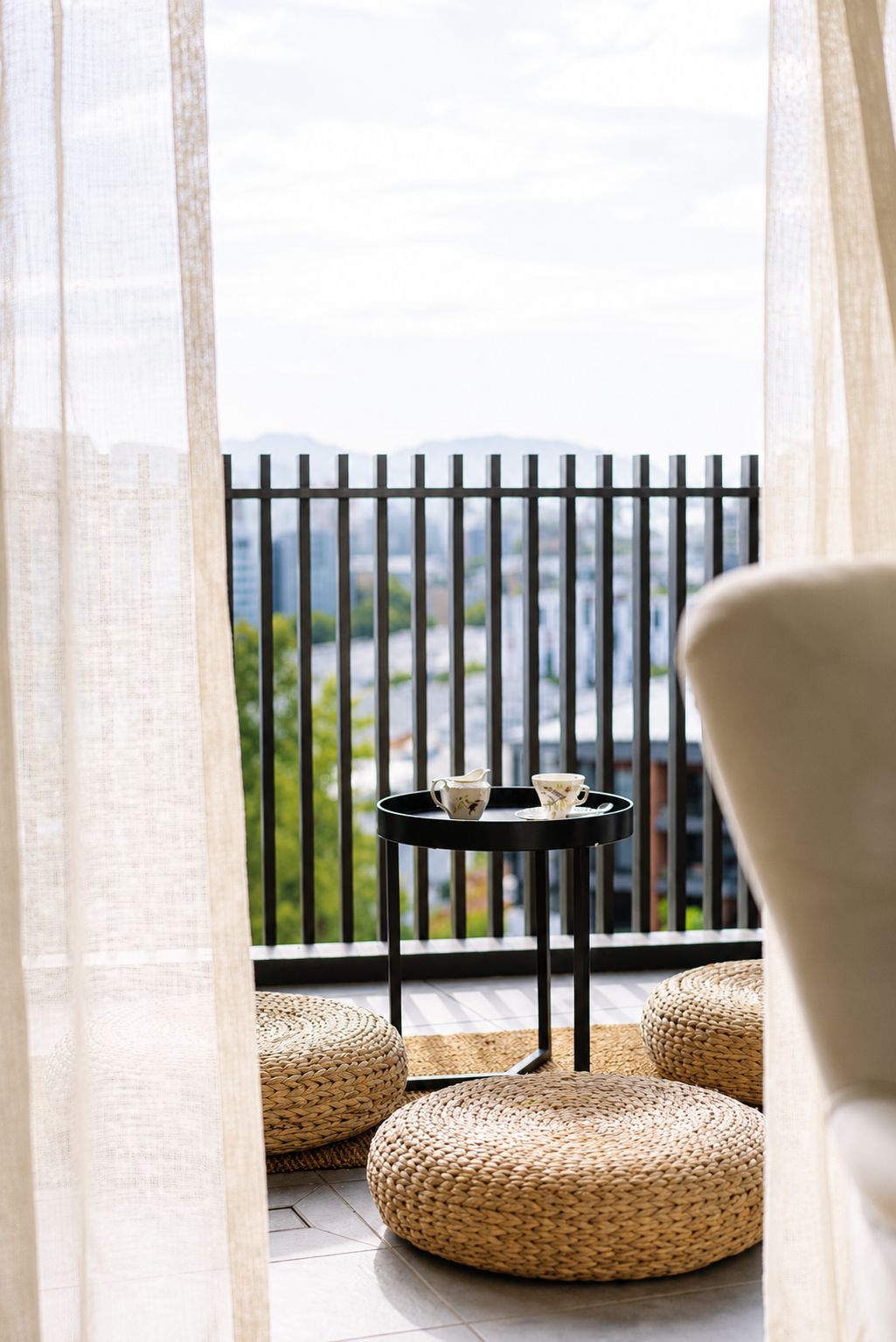 A balcony with a black metal railing, a small round table with two cups, and woven poufs.