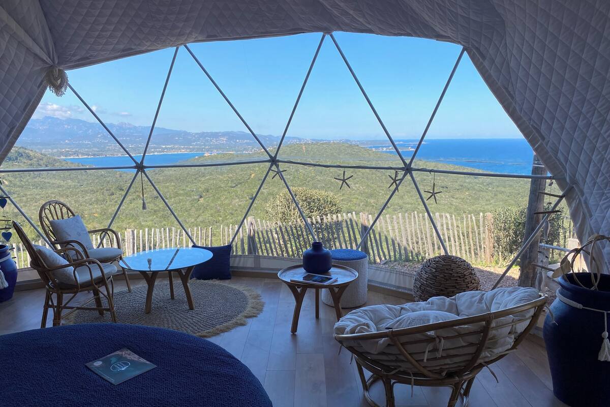 A cozy room with a view of a mountainous landscape and the sea. It includes wicker furniture, a small table, and a comfortable seating area.