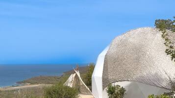 Un edificio de forma abovedada, blanco, con un exterior de fibras naturales trenzadas, ubicado en un paisaje costero con vista despejada al mar.